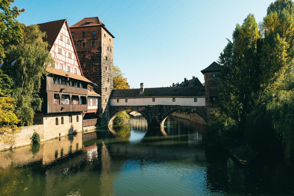 Malerische Ansicht einer Br&uuml;cke in N&uuml;rnberg mit einem historischen Fachwerkhaus und Steinturm, die sich im ruhigen Wasser des Flusses spiegeln, umgeben von gr&uuml;nen B&auml;umen und blauem Himmel.