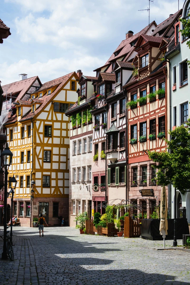 Malerische Stra&szlig;enszene in der Altstadt mit Fachwerkh&auml;usern in verschiedenen Pastellfarben. Kopfsteinpflaster und eine Reihe von historischen Geb&auml;uden mit charakteristischen Dachgauben und Holzfassaden pr&auml;gen das Stra&szlig;enbild. Sonnenlicht und gr&uuml;ne Pflanzen an den Hausfassaden verleihen der Szene eine einladende Atmosph&auml;re.