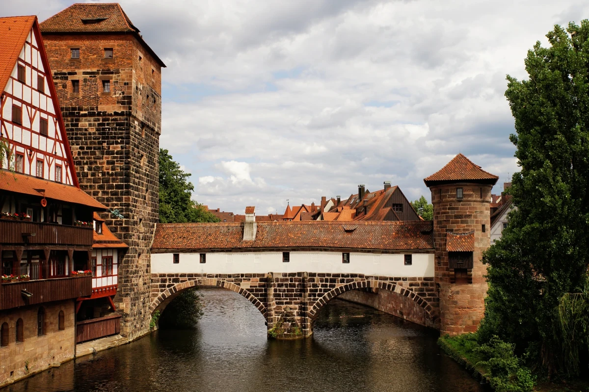 Historische Steinbr&uuml;cke Henkersteg &uuml;ber einen Fluss in N&uuml;rnberg, umgeben von mittelalterlichen Fachwerkh&auml;usern und T&uuml;rmen unter bew&ouml;lktem Himmel, typisch f&uuml;r die architektonische Sch&ouml;nheit der alten Stadt.
