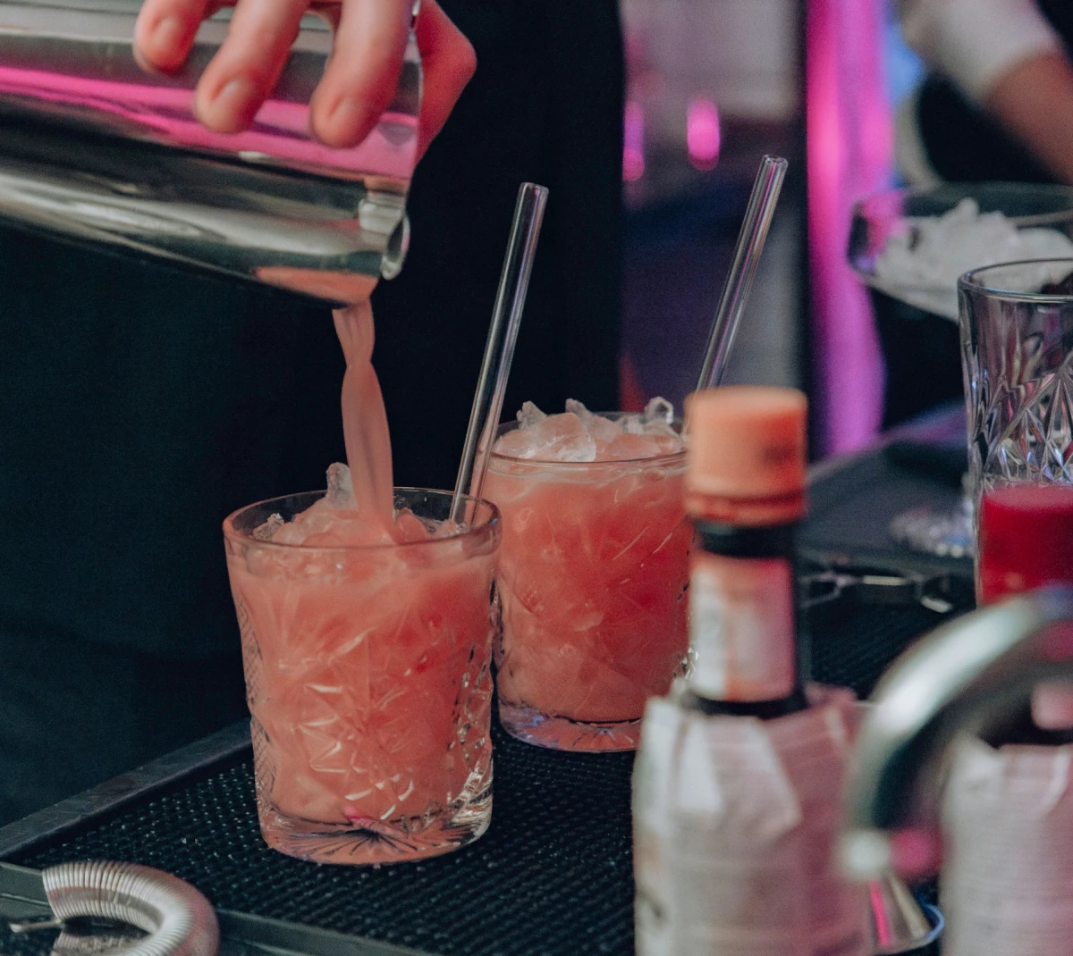Barkeeper gie&szlig;t einen pfirsichfarbenen Cocktail in ein gezacktes Glas mit Eisw&uuml;rfeln, umgeben von Cocktail-Shakern und Barzubeh&ouml;r in ged&auml;mpftem Licht.