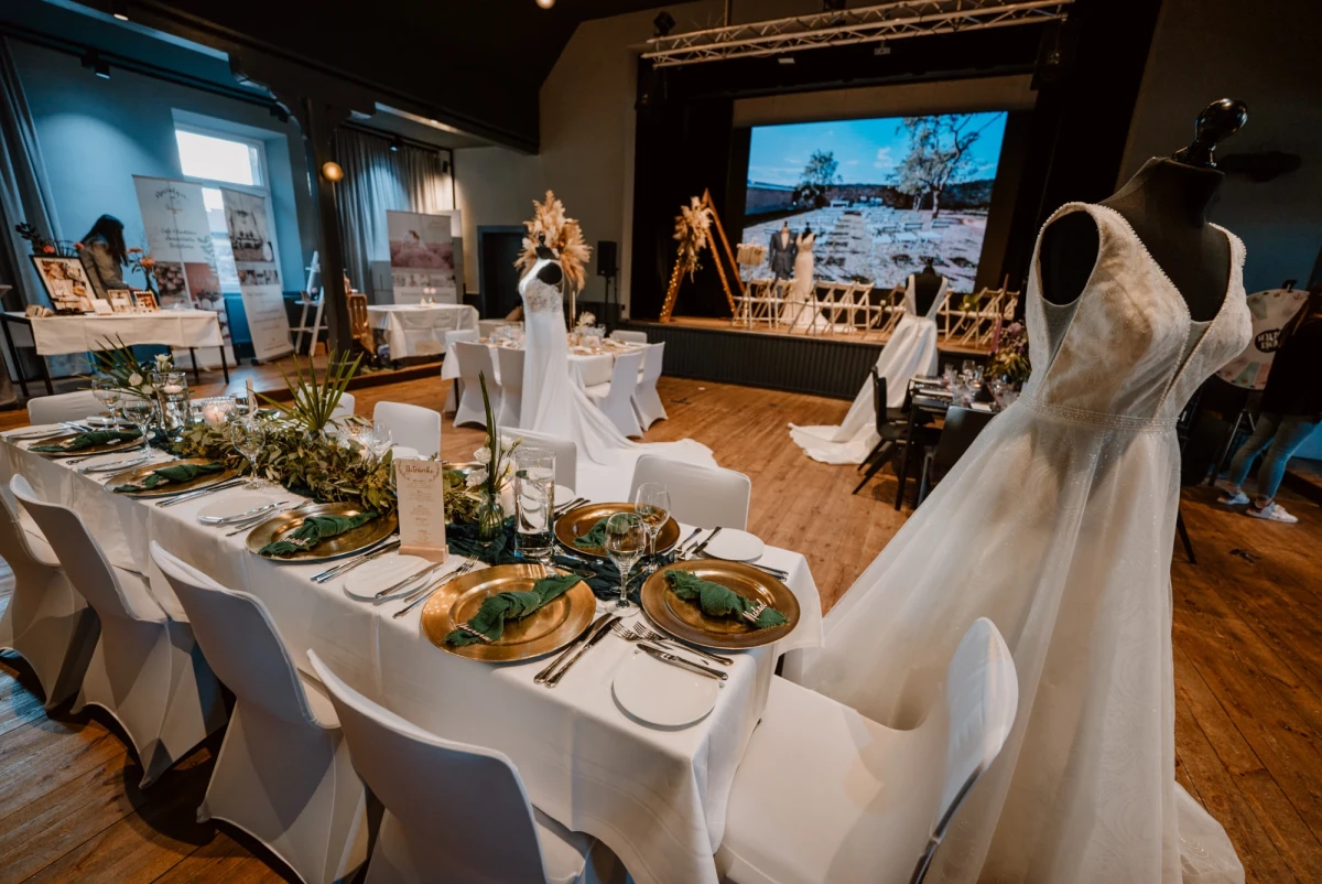 Festlich gedeckter Tisch bei einer Hochzeitsausstellung mit wei&szlig;er Tischdecke, goldenen Tellern und Gr&uuml;ndekorationen. Ein elegantes Brautkleid h&auml;ngt im Vordergrund, im Hintergrund eine Projektionswand mit Landschaftsbild und weitere Hochzeitsdekoration.