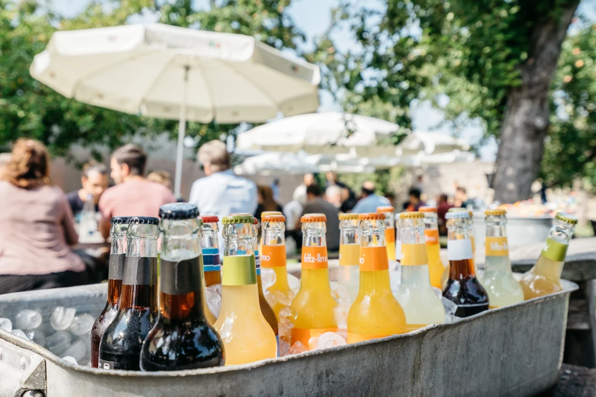 Verschiedene Bierflaschen in unterschiedlichen Farben und Gr&ouml;&szlig;en in einer Metallwanne, im Hintergrund ein wei&szlig;er Sonnenschirm und verschwommene Personen auf einer Sommerparty.