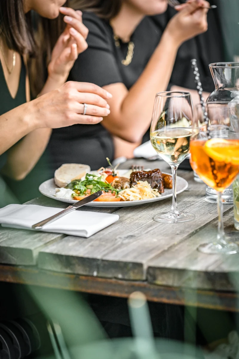 Gedeckter Holztisch mit einem Teller mit Hauptgericht, Salat und Besteck, daneben ein Glas Wei&szlig;wein und ein orangefarbener Cocktail. Personen in dunkler Kleidung sitzen am Tisch und greifen nach Essen und Gl&auml;sern.