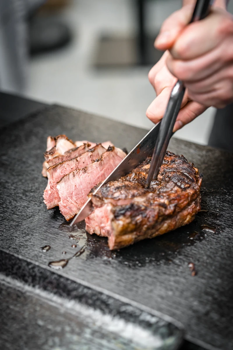 Gebratenes, rosafarbenes Rindfleisch wird mit einer Grillzange auf einer dunklen Steinplatte geschnitten. Das saftige Steak ist medium gegart und hat eine appetitliche, anger&ouml;stete Kruste.