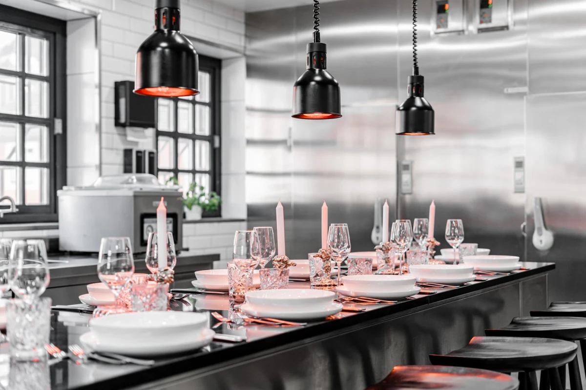 Stilvolle Restaurantk&uuml;che mit schwarzen H&auml;ngelampen &uuml;ber einer Theke. Gedeckter Tisch mit wei&szlig;em Geschirr und Weingl&auml;sern in eleganter, minimalistischer Schwarz-Wei&szlig;-Optik. Edelstahl-K&uuml;chenr&uuml;ckwand und gro&szlig;e Fenster sorgen f&uuml;r einen modernen, hellen Eindruck.