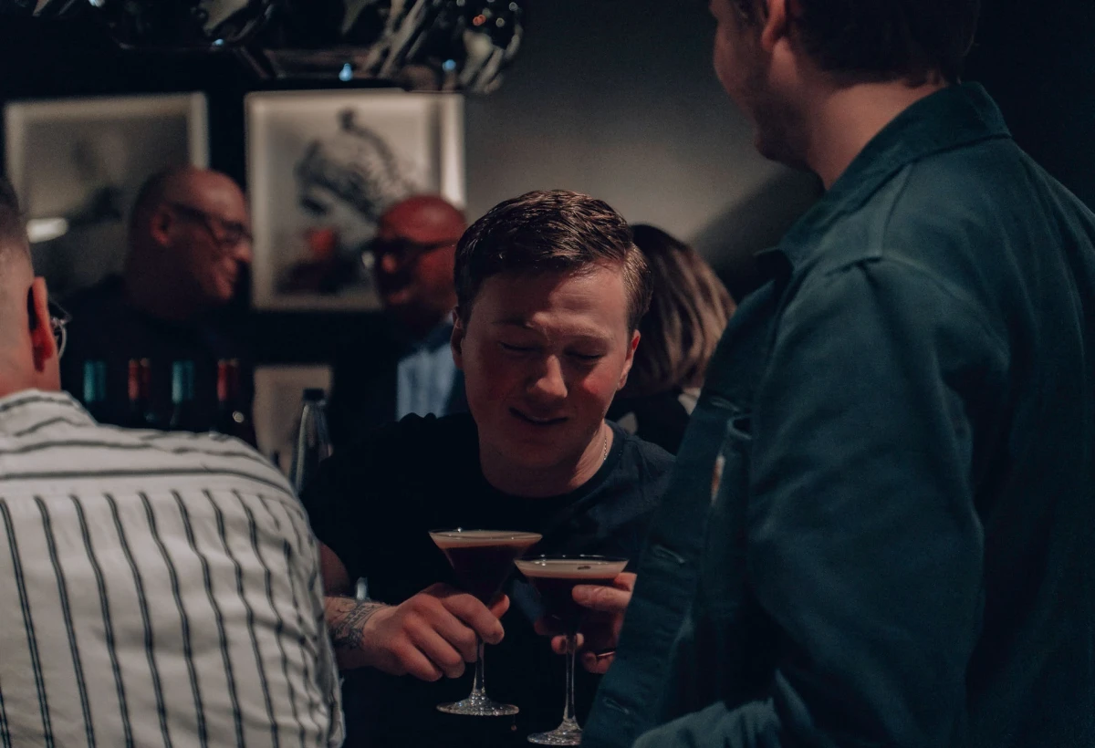 Eine Gruppe von M&auml;nnern an einer Bar, die einen Drink genie&szlig;en. Einer h&auml;lt ein Cocktailglas mit einem dunklen Getr&auml;nk und tr&auml;gt ein gestreiftes Hemd. Die Atmosph&auml;re ist ged&auml;mpft und intim.