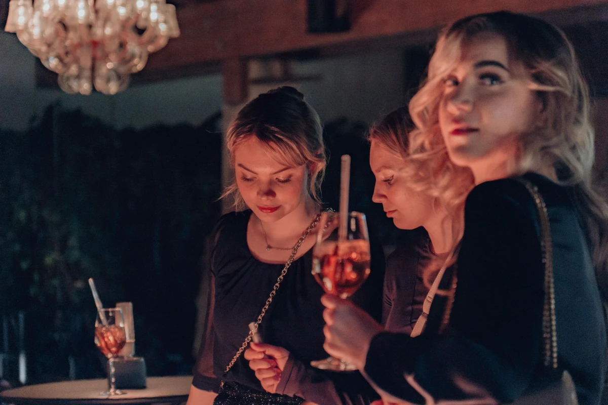 Drei junge Frauen sitzen an einer Bar mit ged&auml;mpftem Licht und einem Kronleuchter im Hintergrund. Sie tragen schwarze Kleidung und halten Cocktails, w&auml;hrend sie in gedankenverlorener Stimmung an der Bar lehnen.