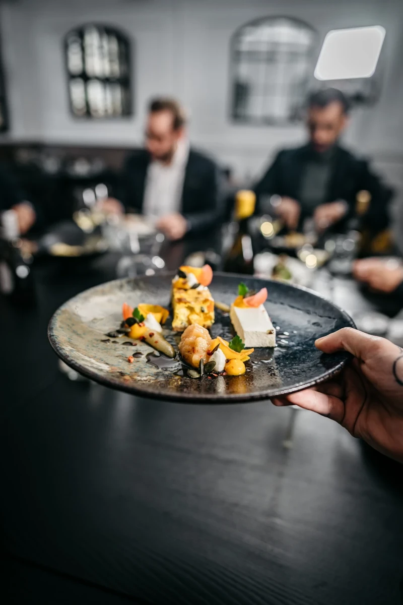 Elegantes Gericht auf einem dunklen Teller mit gelben und orangefarbenen Akzenten, serviert in einem ged&auml;mpften Restaurant mit zwei unscharfen Personen im Hintergrund.