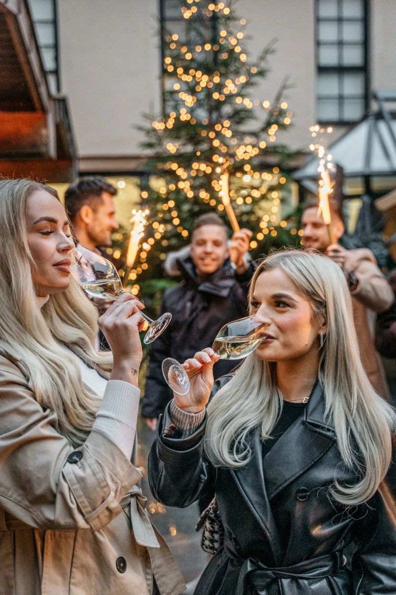 Zwei Frauen mit langen, blonden Haaren trinken Sekt vor einem weihnachtlich beleuchteten Tannenbaum. Sie tragen Winterm&auml;ntel und scheinen eine festliche Feiertagsstimmung zu genie&szlig;en. Im Hintergrund sind weitere Personen in einer gem&uuml;tlichen, weihnachtlichen Atmosph&auml;re zu sehen.