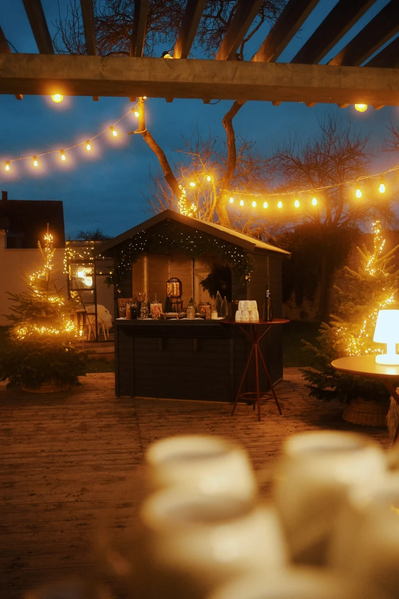 Weihnachtlich beleuchtete Holzh&uuml;tte mit warmen Lichterketten in der Abendd&auml;mmerung, umgeben von Tannenb&auml;umen und einer gem&uuml;tlichen Pergola, im Vordergrund verschwommene Gegenst&auml;nde auf einem Tisch.