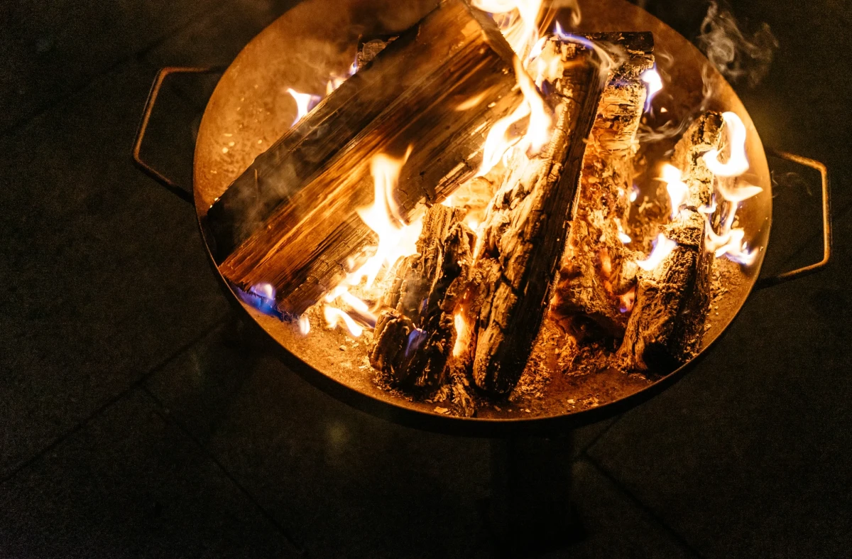 Loderndes Lagerfeuer in einer Feuerschale mit brennenden Holzscheiten, die hell und intensiv in der Dunkelheit leuchten. Die Flammen tanzen um die Holzst&uuml;cke und erzeugen eine warme, einladende Atmosph&auml;re.