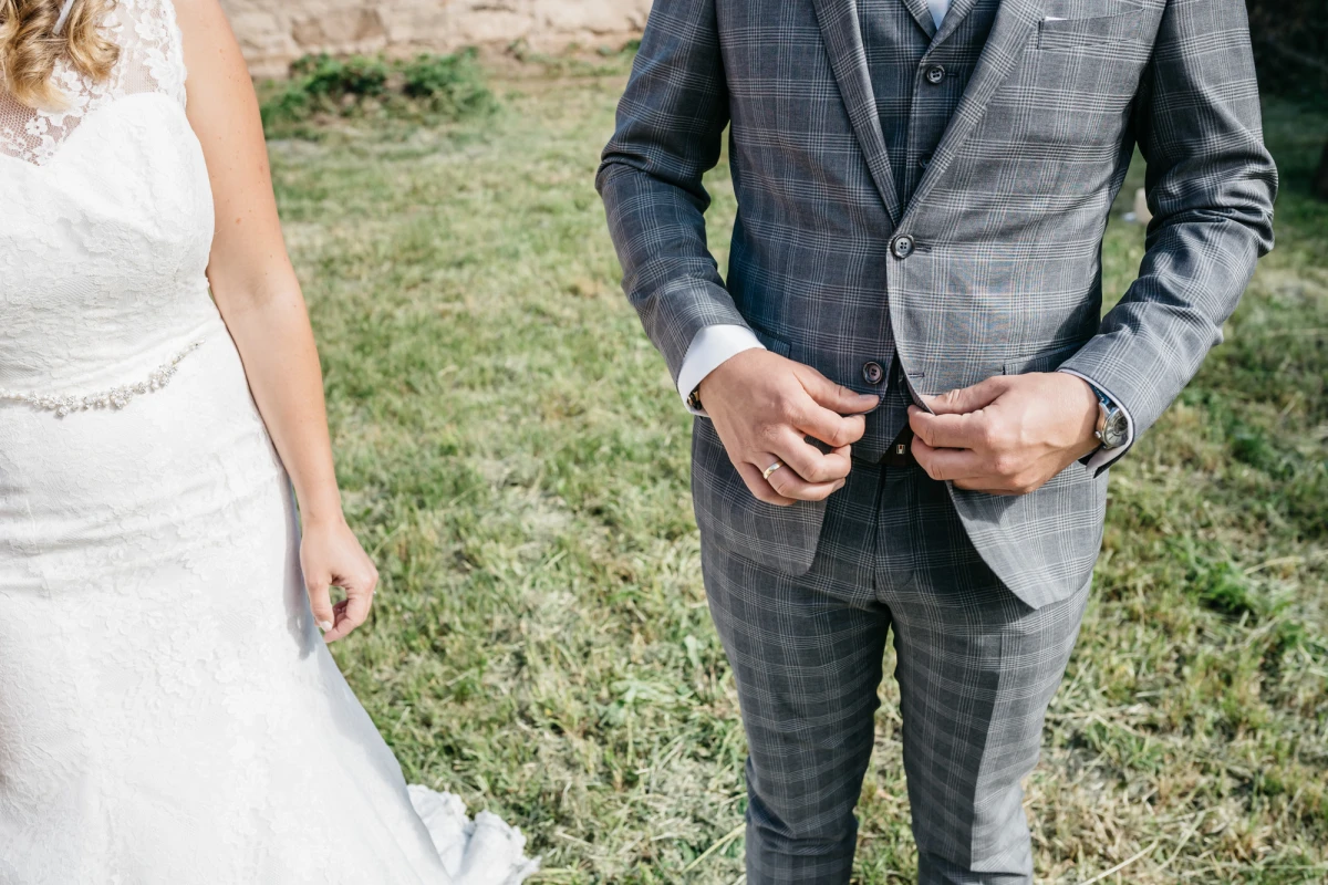 Detailaufnahme einer Hochzeitsszene mit einem Br&auml;utigam in einem grauen karierten Anzug, der seine Jacke schlie&szlig;t, neben einer Braut in einem wei&szlig;en Spitzenkleid auf einer Wiese.