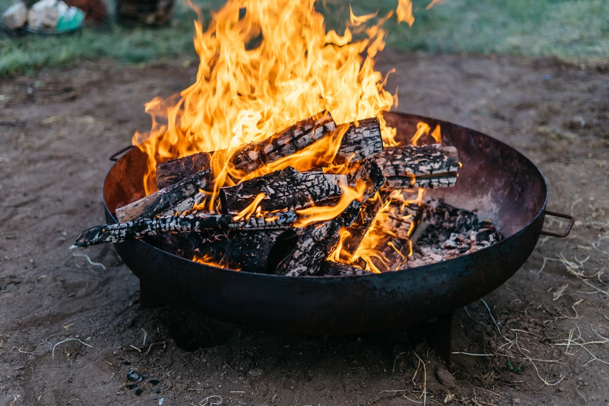 Brennende Holzscheite in einer schwarzen Feuerschale auf dem Boden. Lodernde Flammen in Gelb- und Oranget&ouml;nen umgeben die Holzkohle. Die Glut leuchtet hell vor einem unscharfen Hintergrund.
