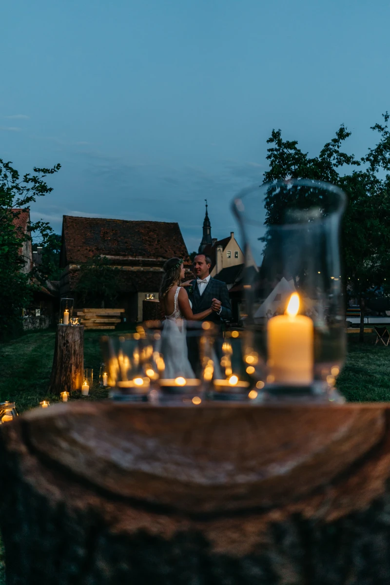 Abendd&auml;mmerung auf einem romantischen Hochzeitsort mit Kerzen im Vordergrund und verschwommenen Brautleuten im Hintergrund. Rustikale Holzscheibe und brennende Kerzen bilden den Rahmen f&uuml;r eine stimmungsvolle Szene mit einer Kirche und B&auml;umen in der D&auml;mmerung.