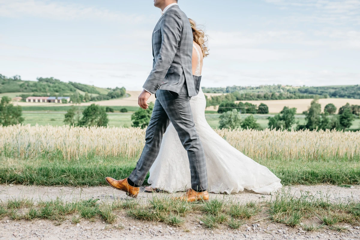 Brautpaar auf einem Feldweg, der Br&auml;utigam in grauem Anzug und gelben Schuhen, die Braut in einem wei&szlig;en Brautkleid, im Hintergrund eine weite Landschaft mit Feldern und B&auml;umen.