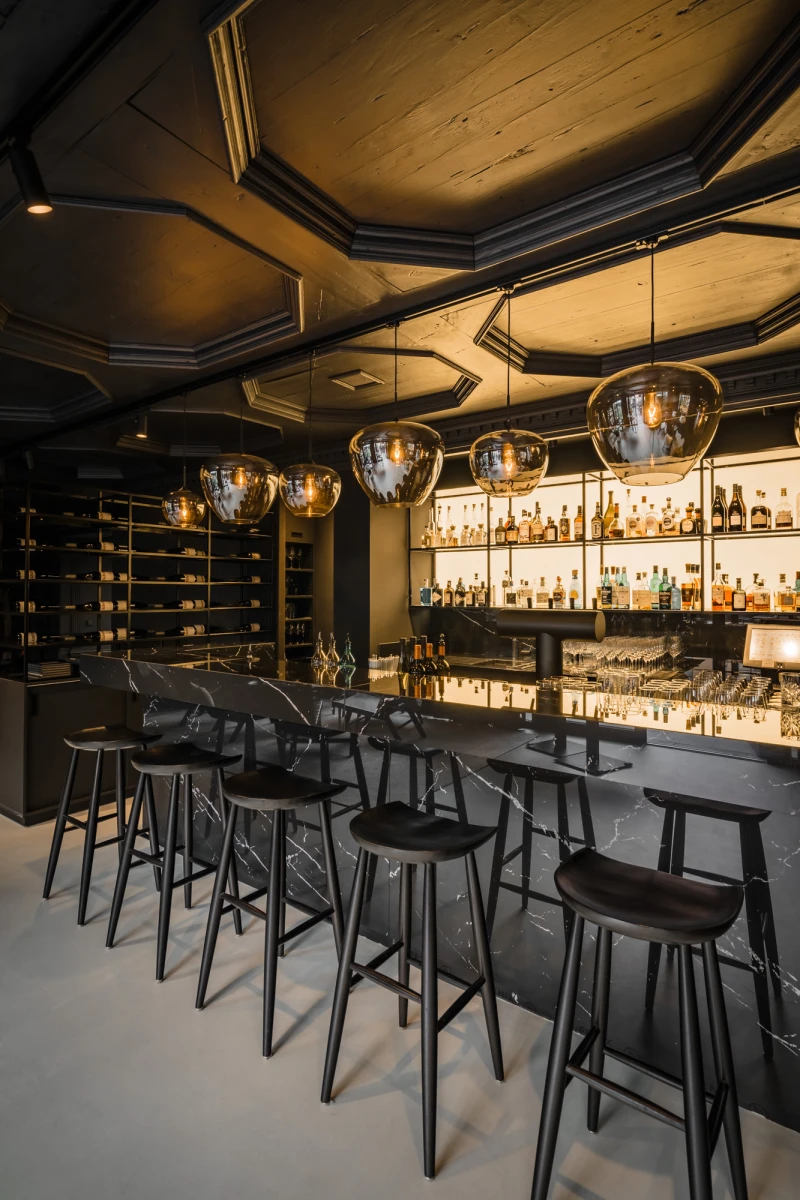 Elegante Bar mit dunklen Holzhockern, die an einer langen Theke stehen. &Uuml;ber der Theke h&auml;ngen gro&szlig;e, runde Pendelleuchten, und die R&uuml;ckwand ist mit beleuchteten Regalen voller Spirituosenflaschen best&uuml;ckt. Die ged&auml;mpfte Beleuchtung und die warmen Holzt&ouml;ne schaffen eine einladende, edle Atmosph&auml;re.