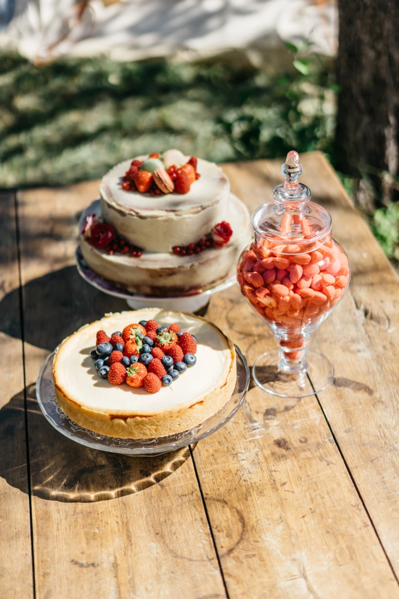 Zwei Obsttorten mit Beeren auf einem rustikalen Holztisch, daneben ein Glas mit roten Erdbeeren. Die Torten sind mit verschiedenen Beerensorten wie Erdbeeren, Blaubeeren und Himbeeren dekoriert, vor einem unscharfen gr&uuml;nen Hintergrund.