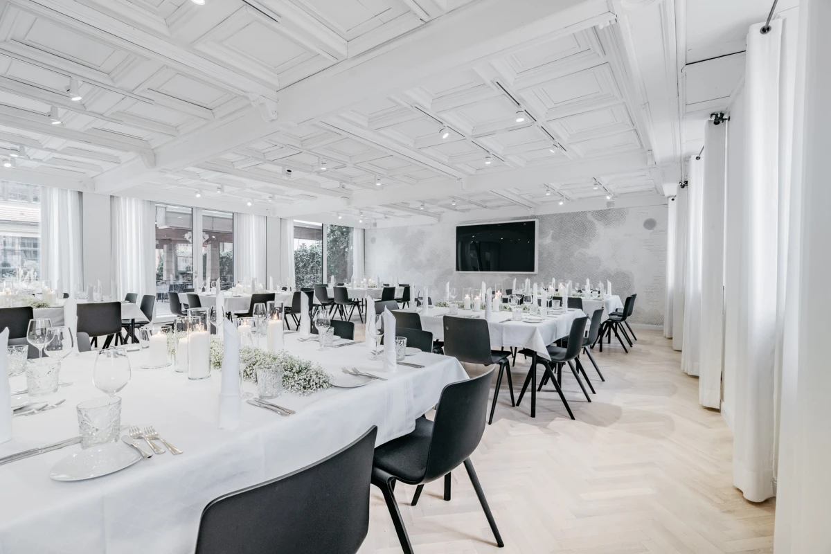 Eleganter Veranstaltungsraum in Wei&szlig; und Schwarz mit gedeckten Tischen, schwarzen St&uuml;hlen und einem gro&szlig;en Fernseher an der Wand. Die Decke ist mit Spotlights ausgestattet und gro&szlig;e Fenster lassen viel nat&uuml;rliches Licht herein, was eine helle und moderne Atmosph&auml;re schafft.