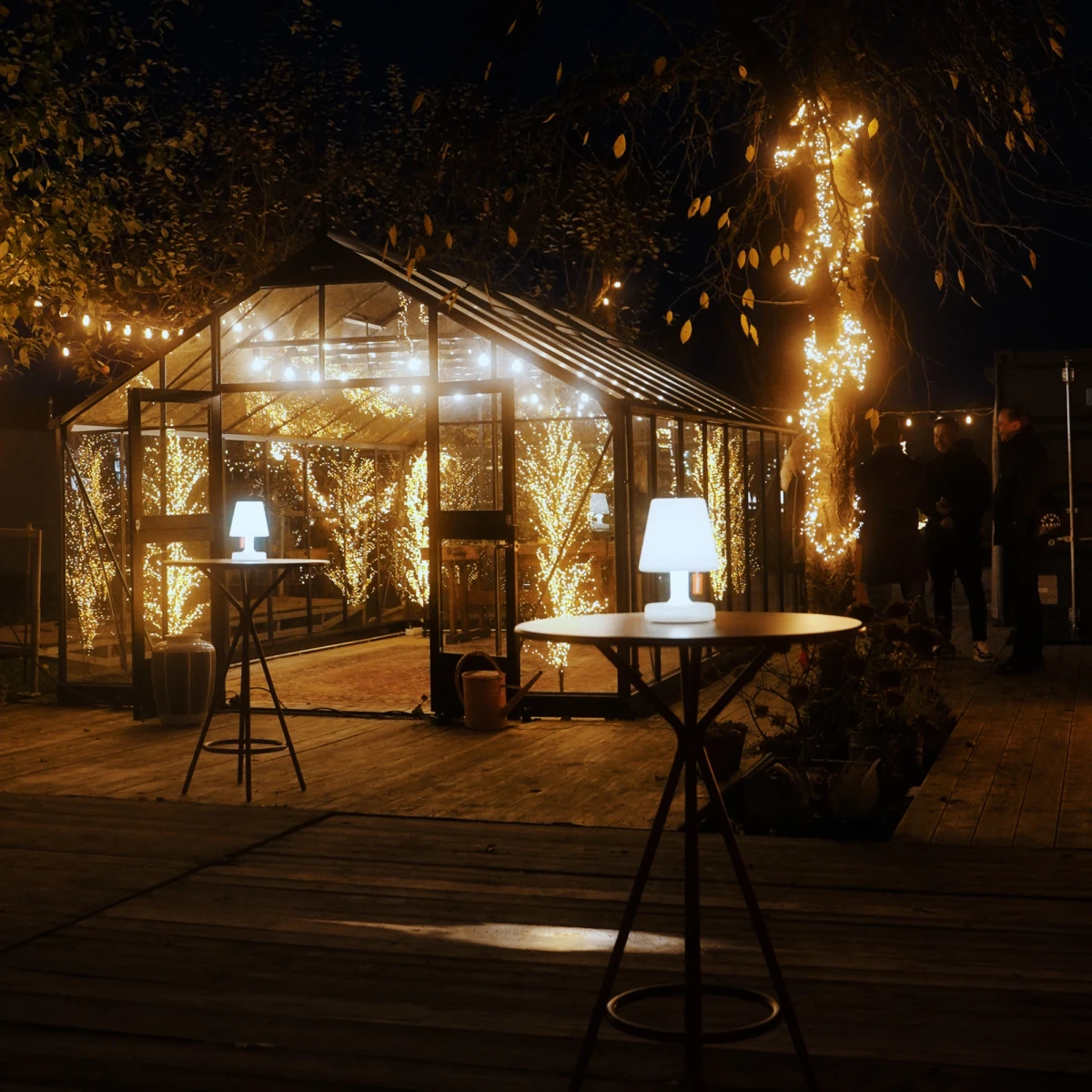 Beleuchtetes Gew&auml;chshaus in der Nacht mit warmen Lichterketten, umgeben von dunklen B&auml;umen. Zwei Stehlampen und Tische im Vordergrund auf einer Holzterrasse, die eine gem&uuml;tliche und romantische Atmosph&auml;re ausstrahlen.