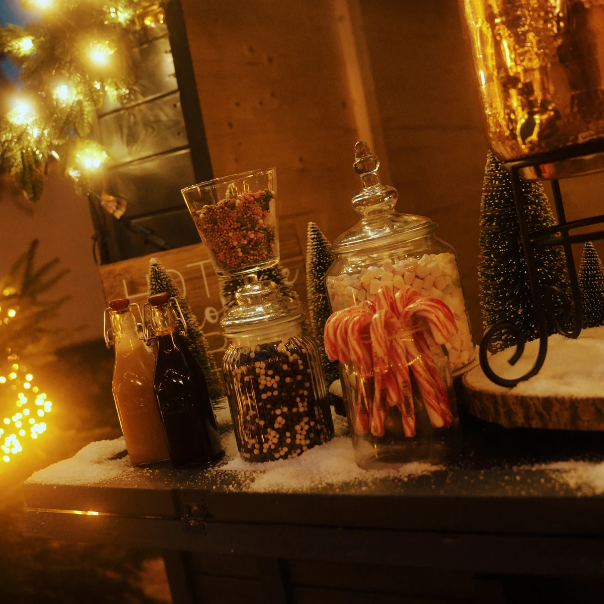 Weihnachtliche Szenerie mit Glasgef&auml;&szlig;en voller S&uuml;&szlig;igkeiten, Lichterketten und Weihnachtsdekoration, die auf schneebedecktem Untergrund in warmem, goldenem Licht gl&auml;nzen.