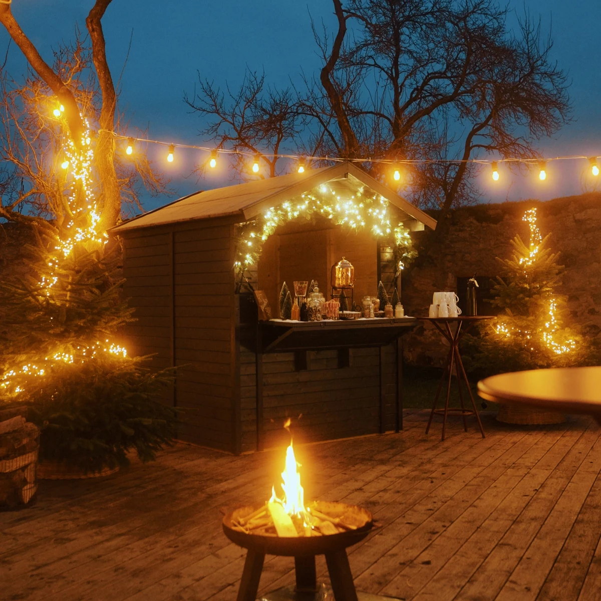 Romantische Winterabendszene mit Lagerfeuer auf einer Holzterrasse, umgeben von beleuchteten B&auml;umen und Lichterketten, mit einer gem&uuml;tlichen Holzh&uuml;tte im Hintergrund, die weihnachtlich geschm&uuml;ckt ist und eine warme, einladende Atmosph&auml;re ausstrahlt.