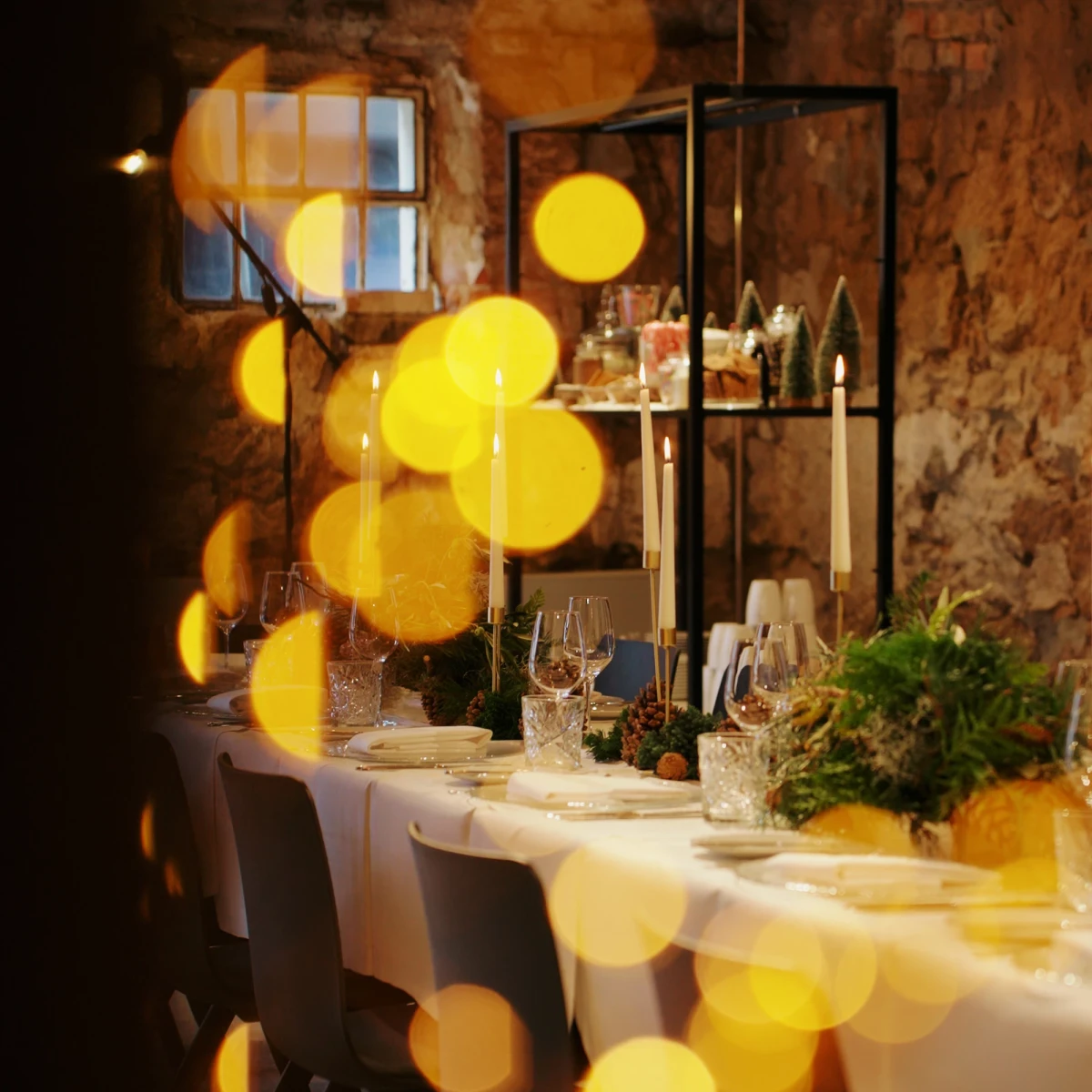 Festlich gedeckter Tisch in rustikaler Steinumgebung mit wei&szlig;er Tischdecke, Gr&uuml;ndekorationen und gelben Lichtreflexen. Weingl&auml;ser und Kerzen auf dem Tisch, im Hintergrund ein Regalgestell und ein Fenster, das warme Atmosph&auml;re vermittelt.