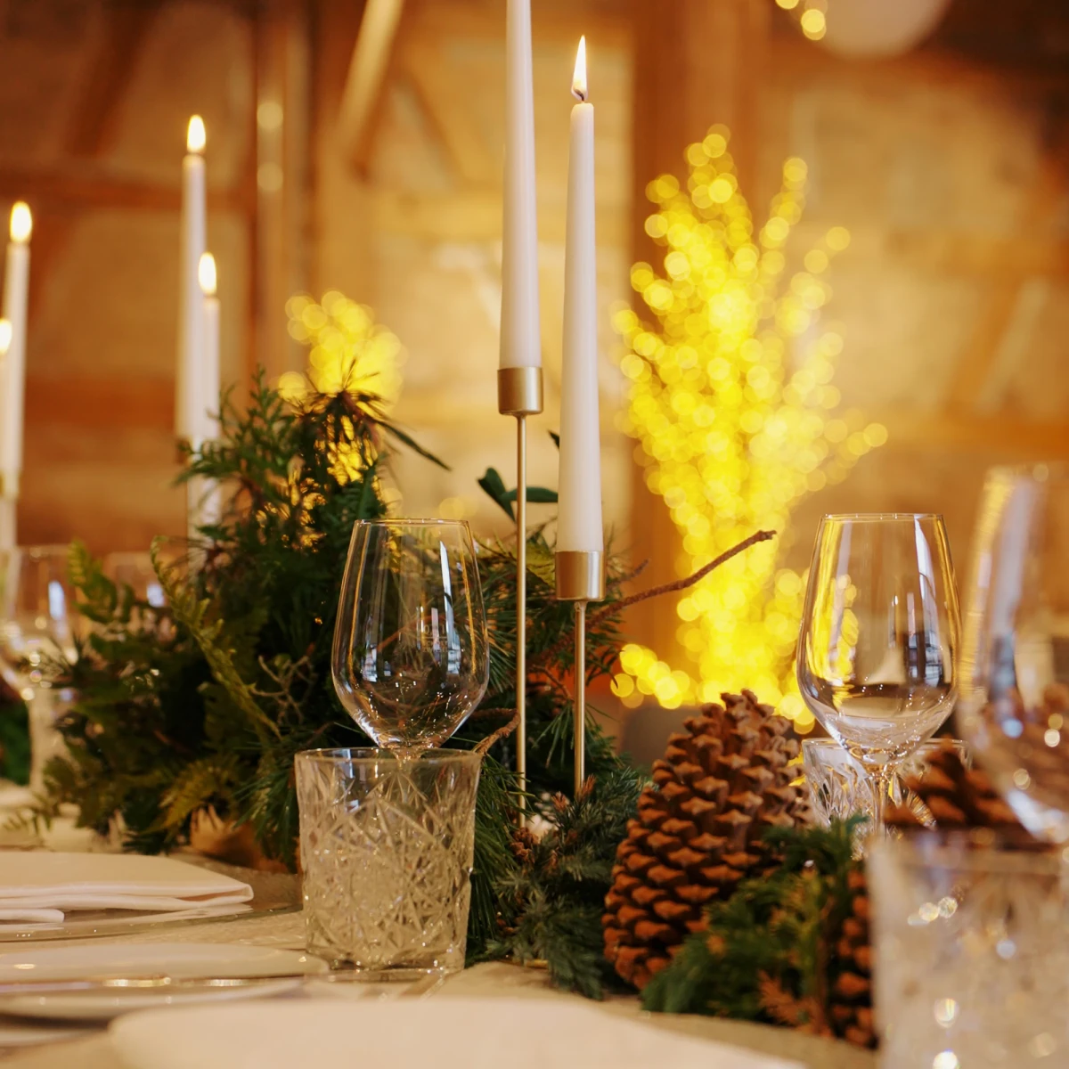 Festlich gedeckter Tisch mit wei&szlig;en Kerzen in Messinghaltern, Weingl&auml;sern und Tannenzweigen, dazu gro&szlig;e Tannenzapfen. Im Hintergrund verschwommene gelbe Lichter, die eine warme, weihnachtliche Atmosph&auml;re erzeugen.