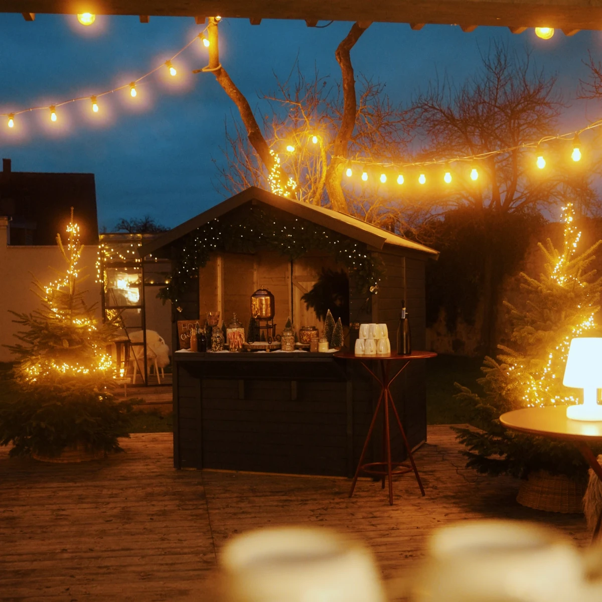 Weihnachtlich beleuchtete Au&szlig;enbar mit warmwei&szlig;en Lichterketten, umgeben von verschneiten B&auml;umen und einer gem&uuml;tlichen Holzh&uuml;tte im Hintergrund, die eine festliche Winterstimmung an einem dunklen Abend einf&auml;ngt.