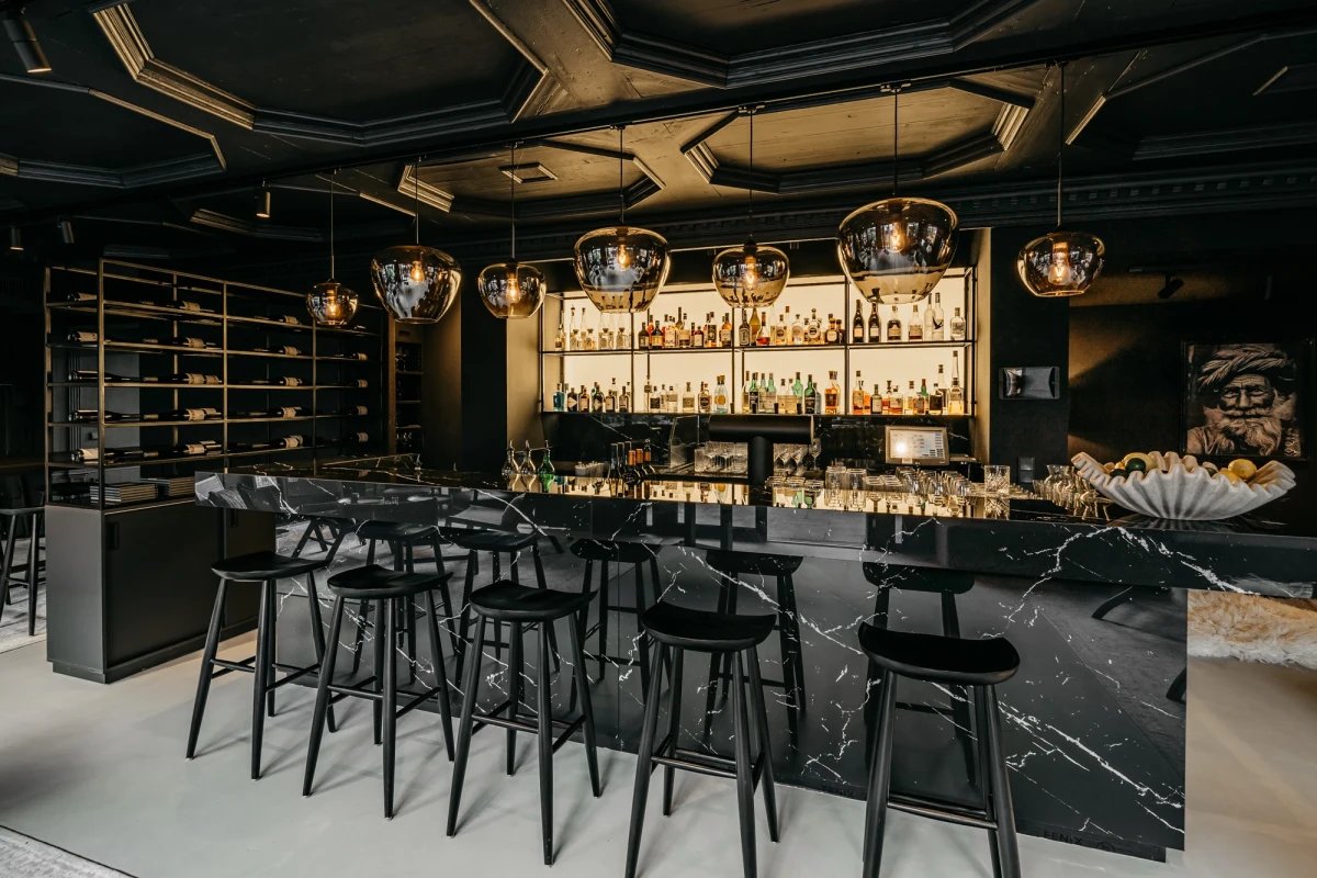 Elegante Bar mit schwarzer Marmorbartheke, schwarzen Barhockern und goldenen H&auml;ngelampen. Dahinter ein beleuchtetes Regal mit zahlreichen Spirituosenflaschen und ein geometrisch strukturierter dunkler Deckenbereich.