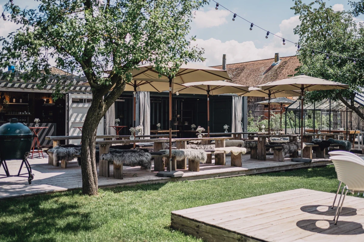 Sonnige Terrasse mit wei&szlig;en Sonnenschirmen, Holzm&ouml;beln und einem Baum, umgeben von gepflegtem Rasen und rustikalen Geb&auml;uden im Hintergrund, perfekt f&uuml;r entspannte Mahlzeiten im Freien.