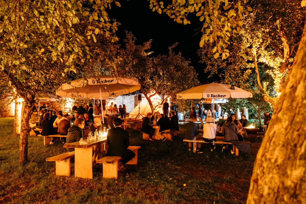Abendliche Gartenszene mit beleuchteten Pavillons und Stehtischen, Menschen versammeln sich unter herbstlichen B&auml;umen mit warmgoldenem Licht und ged&auml;mpfter Atmosph&auml;re.