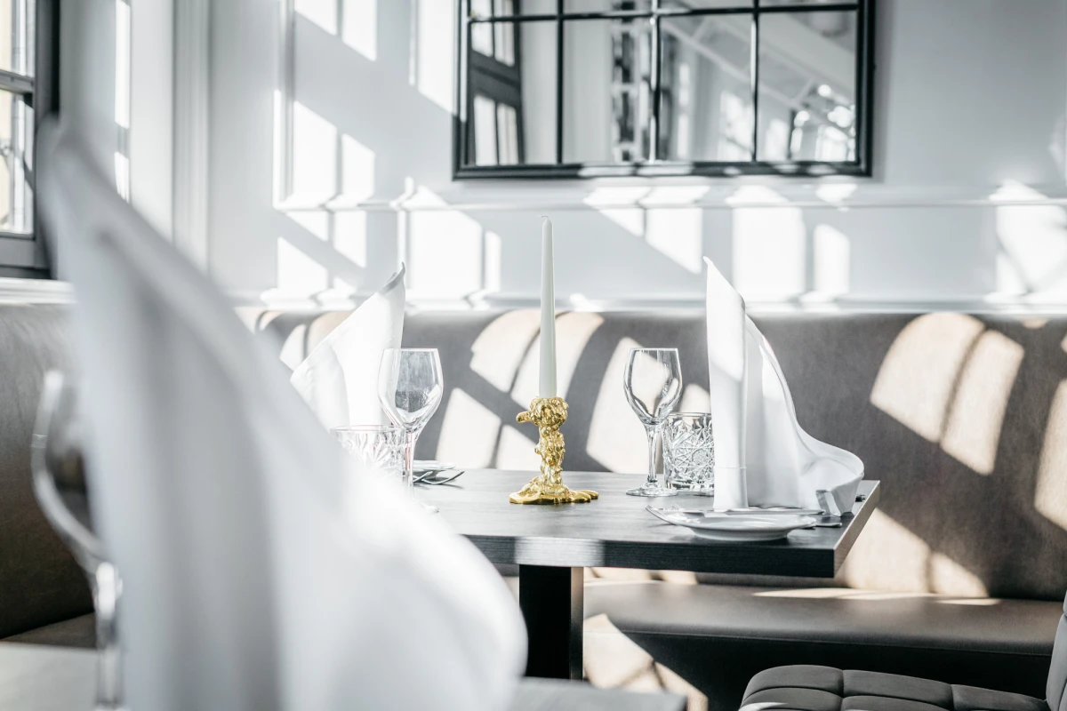 Elegante Restauranttische mit wei&szlig;en Tellern, Weingl&auml;sern und einer goldenen Kerze, durch gro&szlig;e Fenster mit Sonnenlicht durchflutet. Ged&auml;mpfte Atmosph&auml;re mit wei&szlig;en St&uuml;hlen und einem Spiegel an der Wand im Hintergrund.