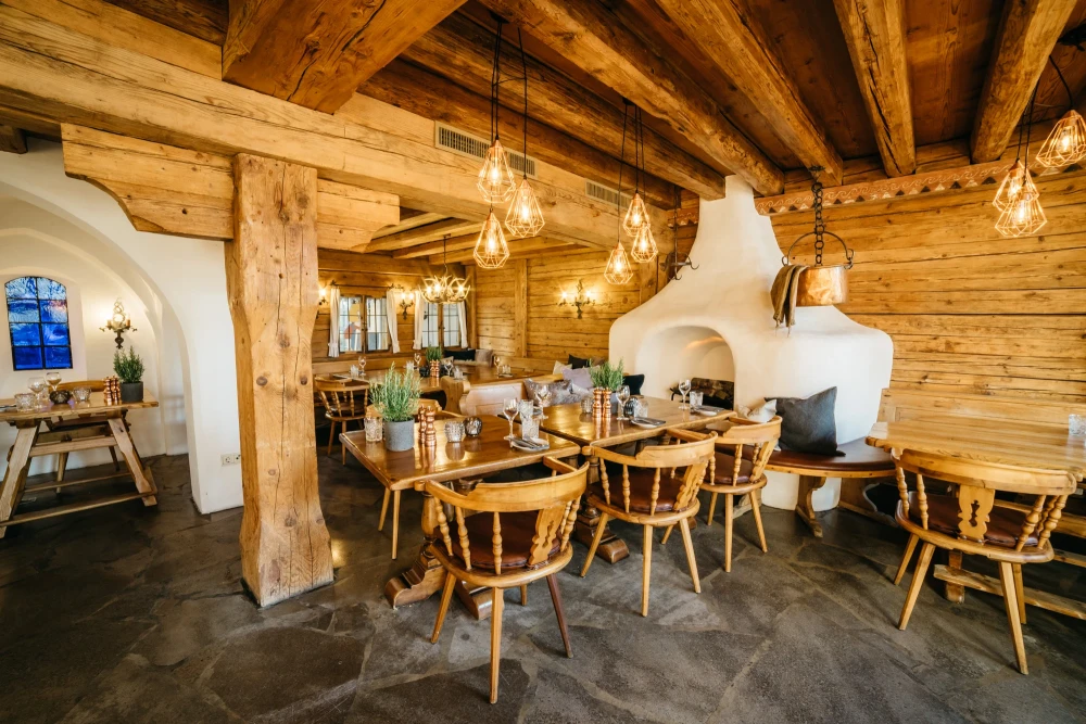 Gem&uuml;tlicher Gastraum in Holzblockhaus-Optik mit rustikalen Holzbalken, h&auml;ngenden Pendelleuchten und gedeckten Holztischen mit St&uuml;hlen. Ein wei&szlig; gefliester Kachelofen und Holzw&auml;nde schaffen eine warme, einladende Atmosph&auml;re in traditionellem Bergstil.