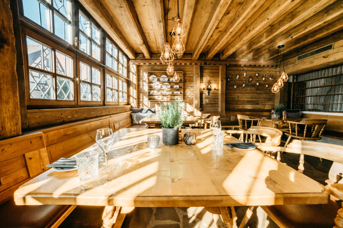 Rustikaler Gastraum mit Holzbalkendecke und gro&szlig;en Fenstern, an einem langen Holztisch gedeckt mit Gl&auml;sern und einer Zimmerpflanze. Vintage H&auml;ngelampen und warmes Sonnenlicht schaffen eine gem&uuml;tliche Atmosph&auml;re im Landhausstil.