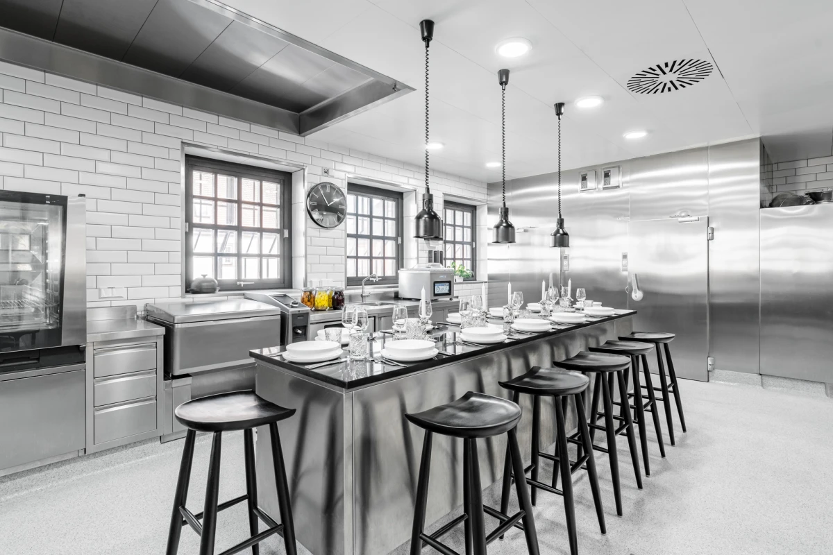 Moderne, gro&szlig;z&uuml;gige Restaurantk&uuml;che mit langer Theke, Barhockern und Edelstahloberfl&auml;chen. Helle, ger&auml;umige K&uuml;cheneinrichtung mit Pendelleuchten und gro&szlig;en Fenstern, die eine professionelle Arbeitsatmosph&auml;re ausstrahlt.