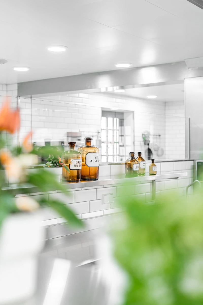Bernsteinfarbene Apothekergl&auml;ser stehen in einer wei&szlig;en K&uuml;che mit wei&szlig;en Wandfliesen und Deckenbeleuchtung. Im Vordergrund verschwimmen gr&uuml;ne Bl&auml;tter und Blumen, w&auml;hrend die Glasflaschen scharf im Fokus sind. Die helle, moderne K&uuml;cheneinrichtung strahlt Sauberkeit und Eleganz aus.