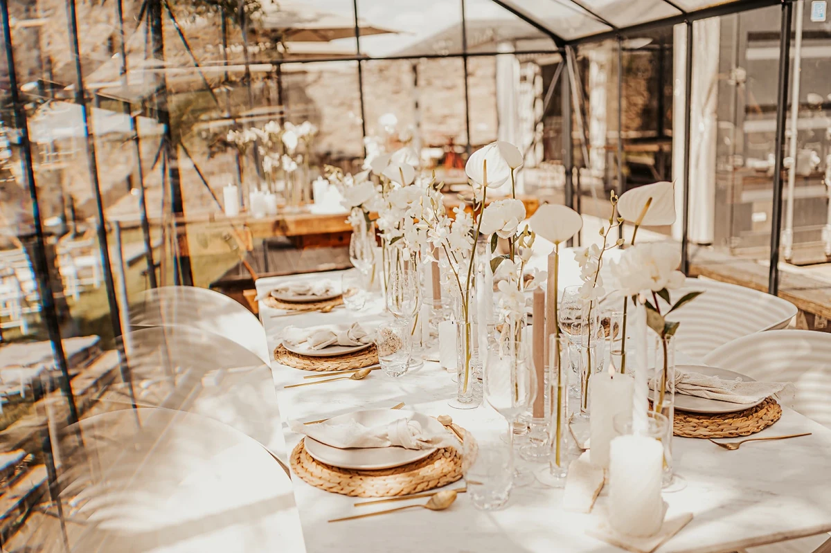 Gedeckter Tisch in einem Wintergarten oder Gew&auml;chshaus mit wei&szlig;er Tischdecke, goldenen Platztellern und eleganten Glasvasen mit wei&szlig;en Blumen. Die Tischdekoration ist minimalistisch und zeitlos, mit einer warmen, einladenden Atmosph&auml;re.