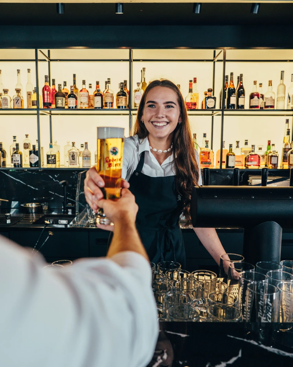 Eine l&auml;chelnde Barkeeperin in Sch&uuml;rze reicht ein Bierglas &uuml;ber die Theke, im Hintergrund eine Reihe von Spirituosenflaschen in einem Bar-Regal.