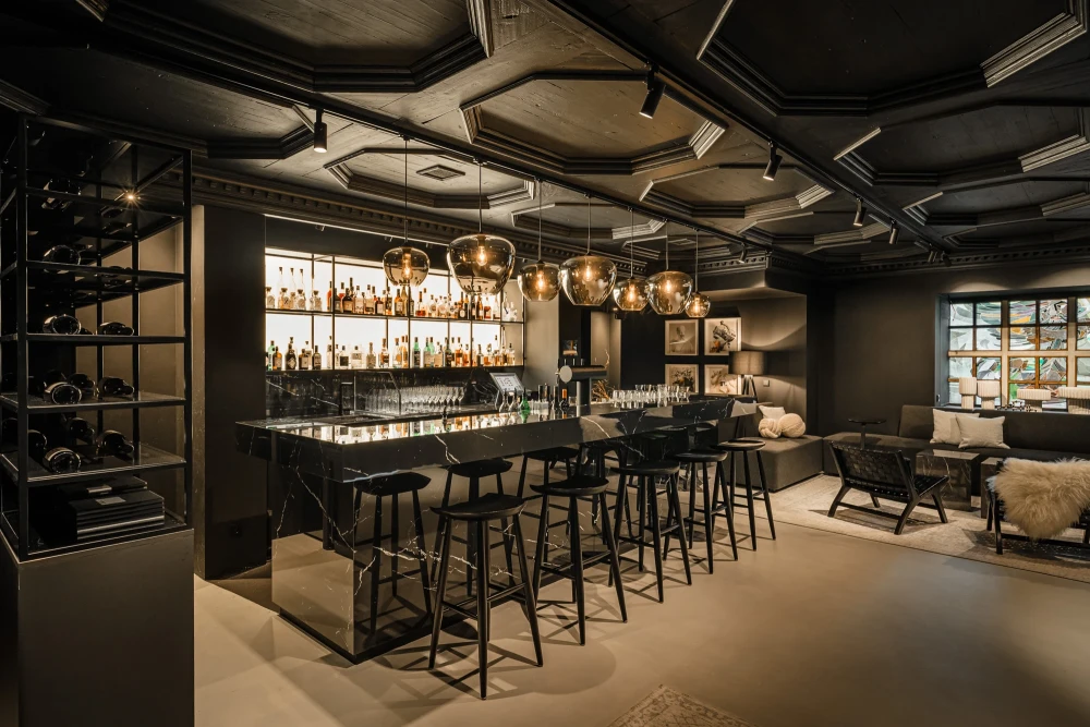 Moderner Bar-Bereich mit schwarzen Barhockern, goldenen Pendelleuchten und einer Regalwand voller Spirituosen. Die Decke ist geometrisch gestaltet, und im Hintergrund ist eine gem&uuml;tliche Sitzecke mit einem Sessel zu sehen.
