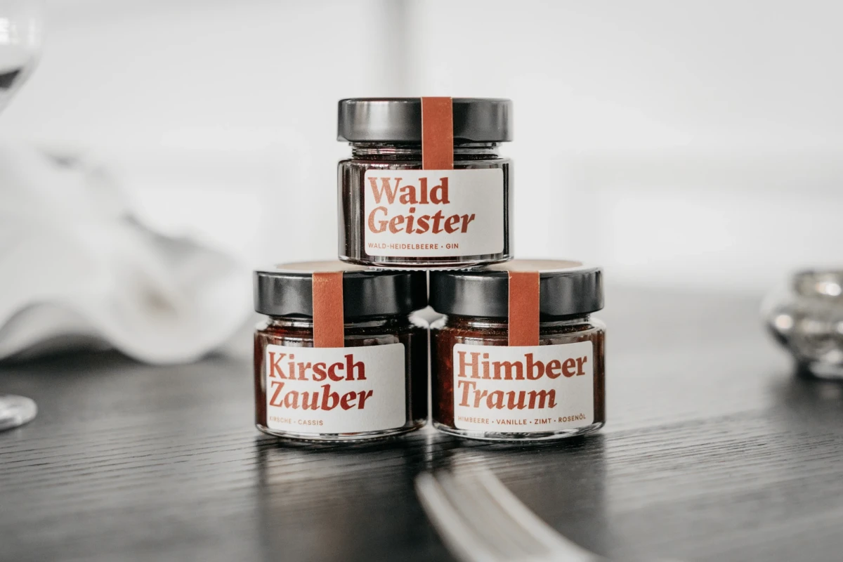 Drei Glasgef&auml;&szlig;e mit verschiedenen Etiketten von Wald Geister, Kirsch Zauber und Himbeer Traum, gestapelt auf einer dunklen Oberfl&auml;che in einem minimalistischen Setting.