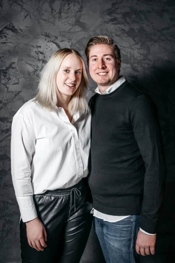 Zwei l&auml;chelnde Personen vor einer grauen Betonwand. Eine Frau in wei&szlig;er Bluse und Lederhose steht links, ein Mann in schwarzem Pullover und blauer Jeans rechts von ihr. Beide stehen eng beieinander und wirken freundlich.