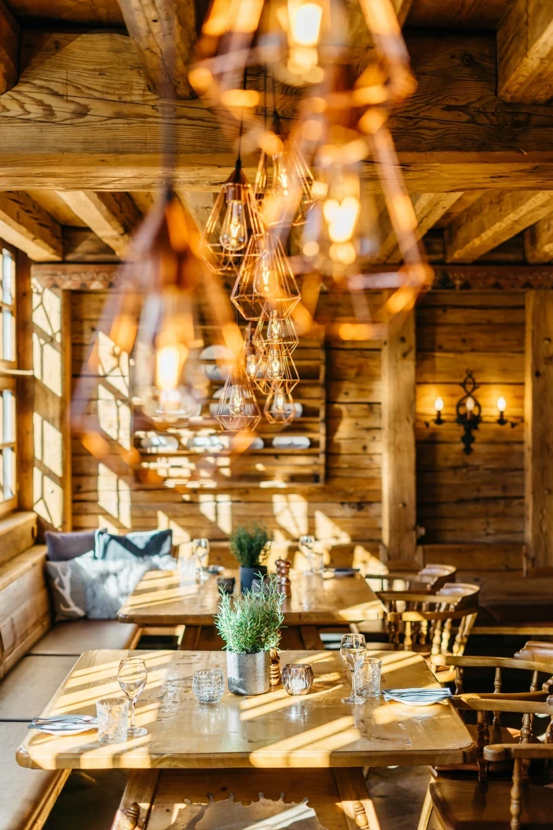 Rustikal eingerichteter Gastraum mit Holzw&auml;nden und -balken, warm beleuchtet durch h&auml;ngende Lampen und Wandleuchten. Gedeckte Holztische mit Blumen in Metallvasen, gem&uuml;tliche Sitzecke mit Kissen und Sonnenlicht, das durch Holzfenster f&auml;llt.