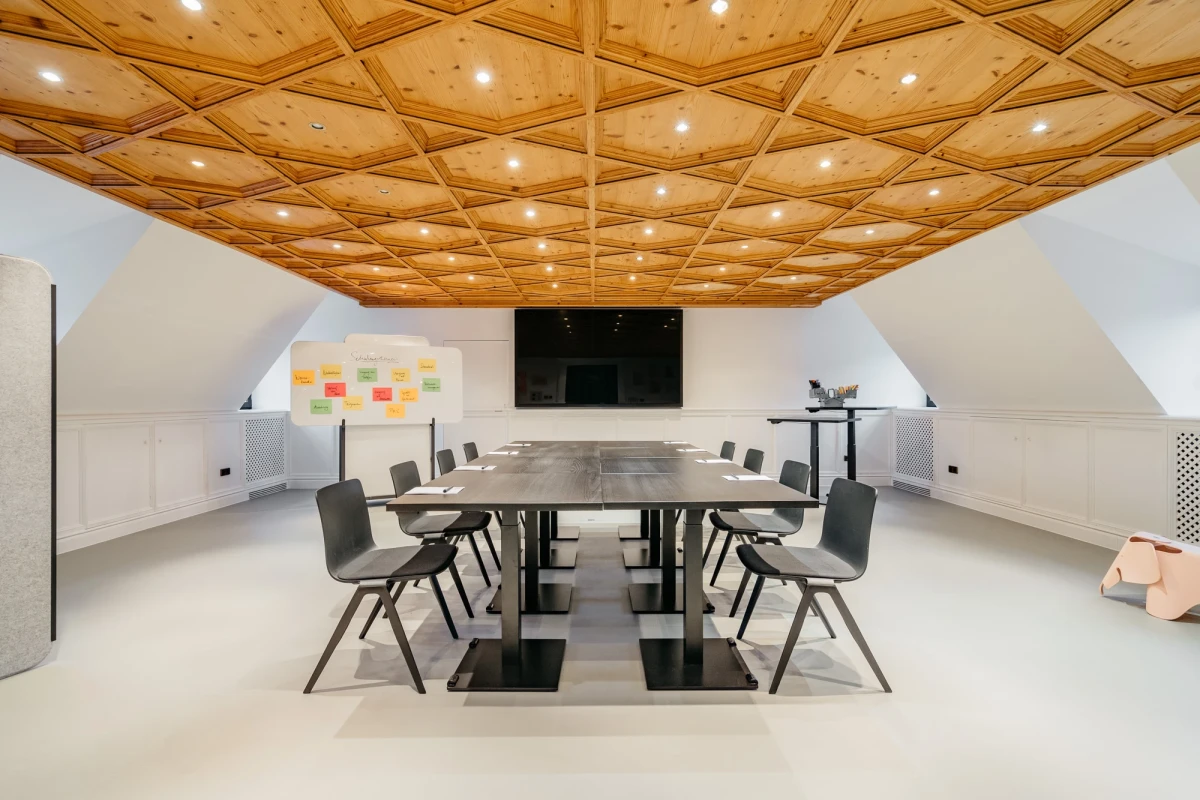 Moderner Konferenzraum mit Holzdecke und geometrischem Muster, ausgestattet mit einem gro&szlig;en Tisch, grauen St&uuml;hlen und einem schwarzen Bildschirm an der Wand. Helle, minimalistisch gestaltete R&auml;umlichkeit mit indirekter Beleuchtung und einer Pinnwand mit bunten Haftnotizen.