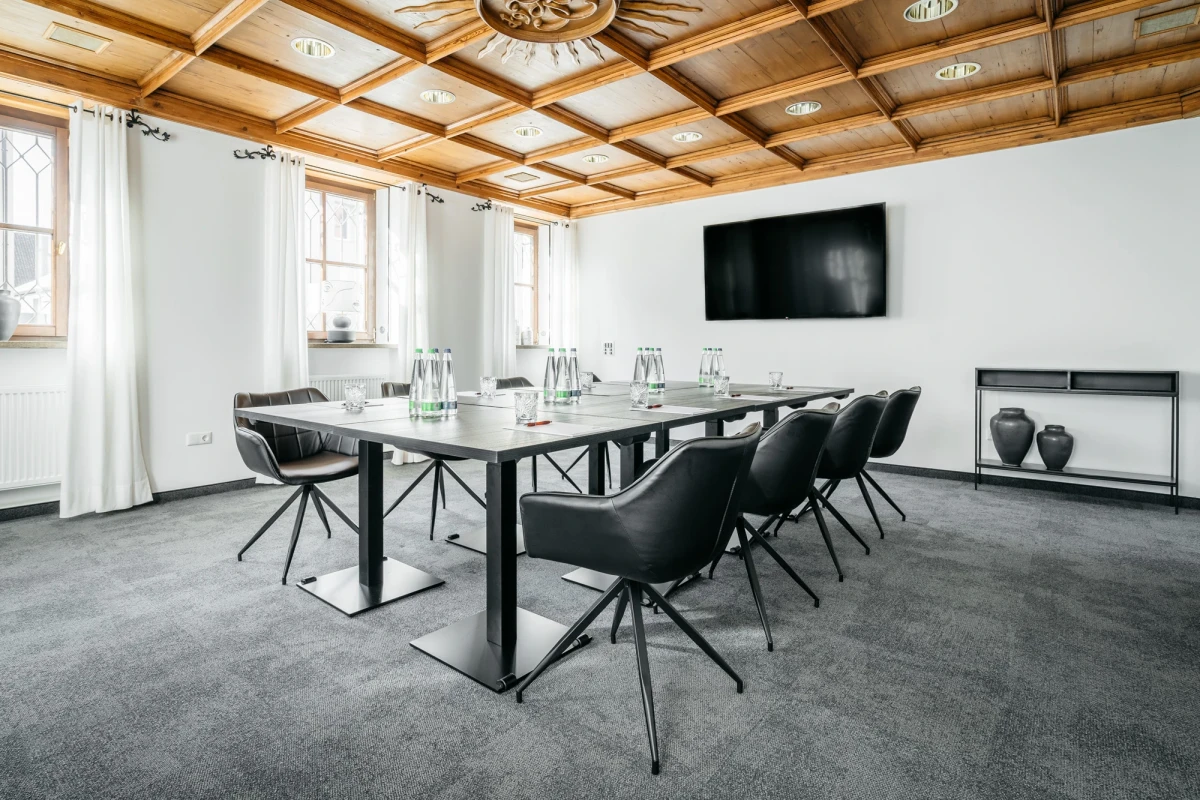 Moderner Konferenzraum mit wei&szlig;en W&auml;nden, holzget&auml;felter Decke und grauem Teppichboden. Ein gro&szlig;er rechteckischer Konferenztisch mit schwarzen Designerst&uuml;hlen steht im Raum, an der Wand h&auml;ngt ein gro&szlig;er schwarzer Fernseher. Die Fenster sind mit wei&szlig;en Vorh&auml;ngen verhangen.