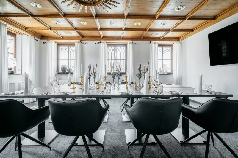 Eleganter Konferenzraum mit gro&szlig;en Fenstern, wei&szlig;er Wandgestaltung und holzget&auml;felter Decke. Langer Tisch mit schwarzen Drehst&uuml;hlen und festlicher Tischdekoration mit goldenen Kerzenhaltern. Modernes, helles Ambiente f&uuml;r Meetings und Veranstaltungen.