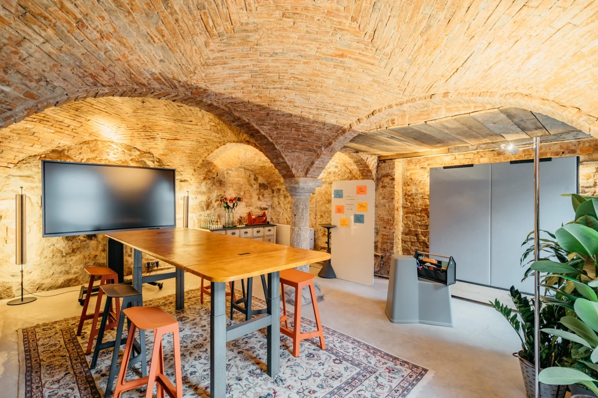 Moderner Besprechungsraum mit Backsteingew&ouml;lbe und historischer Architektur. Ein gro&szlig;er Holztisch mit Barhockern steht auf einem gemusterten Teppich, daneben ein Flachbildschirm und eine Glastrennwand. Warmes Licht und rustikale Steinw&auml;nde schaffen eine einladende Atmosph&auml;re f&uuml;r Meetings.