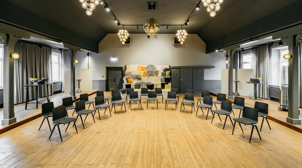 Heller Seminarraum mit Holzboden und modernen schwarzen St&uuml;hlen, die in einem Halbkreis angeordnet sind. Warme Beleuchtung durch Kugelleuchten und ein abstraktes Kunstwerk an der Wand im Hintergrund. Die Atmosph&auml;re wirkt einladend und professionell f&uuml;r Meetings oder Workshops.