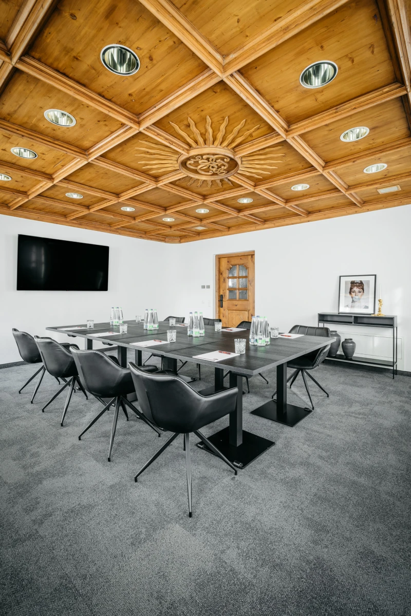 Moderner Konferenzraum mit Holzdeckengestaltung, gro&szlig;em Glastisch und schwarzen Drehst&uuml;hlen. Schwarze Wandtafel und graue Teppichbodengestaltung vervollst&auml;ndigen den eleganten Raum.