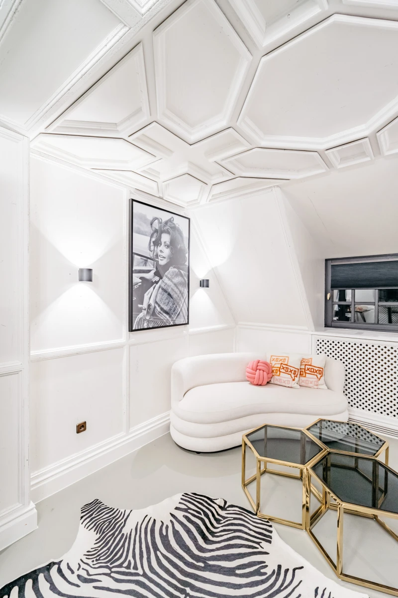 Moderner, wei&szlig;er Wohnraum mit geschwungener Couch und geometrischer Deckengestaltung. Schwarzwei&szlig;es Kunstwerk an der Wand, Zebra-gemusterter Teppich und goldene Beistelltische bilden einen eleganten, minimalistischen Raum.