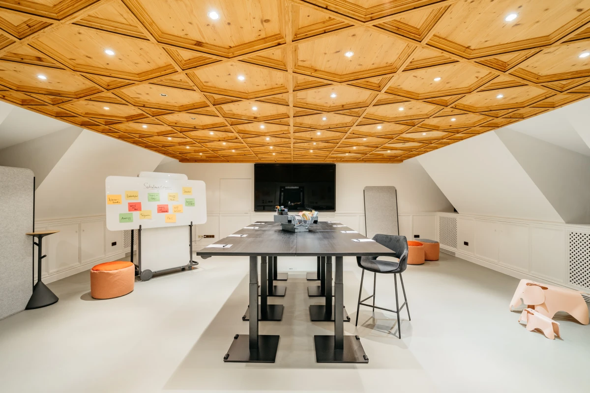 Moderner Besprechungsraum mit geometrischer Holzdecke und Designerbeleuchtung. Ein gro&szlig;er Konferenztisch mit St&uuml;hlen steht in der Mitte, daneben eine Pinnwand und ein Whiteboard. Der Raum wirkt hell, minimalistisch und einladend, mit wei&szlig;en W&auml;nden und warmen Holzt&ouml;nen.